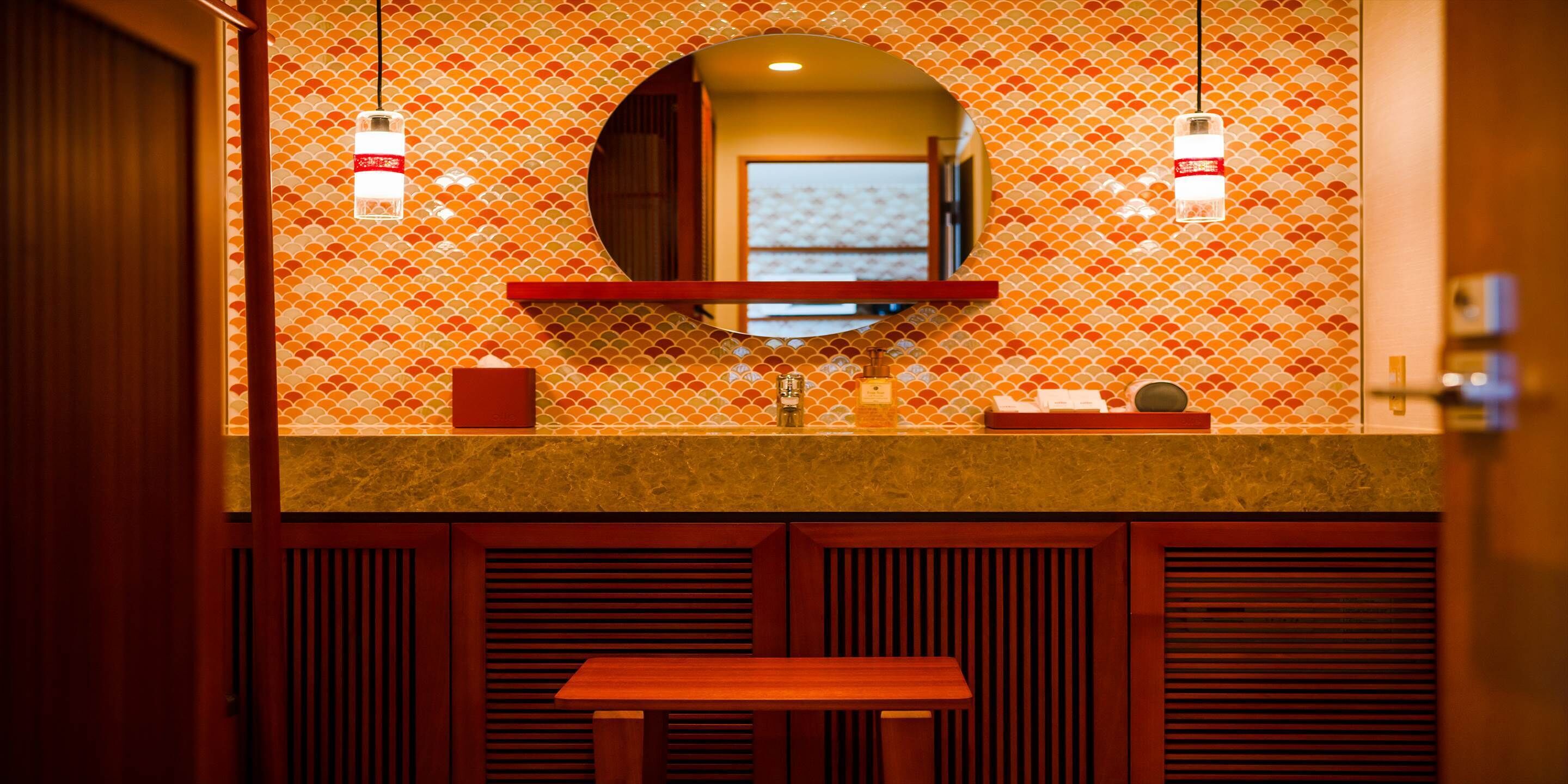 Um banheiro com parede revestida de azulejos, um espelho, uma pia e um banco de madeira.