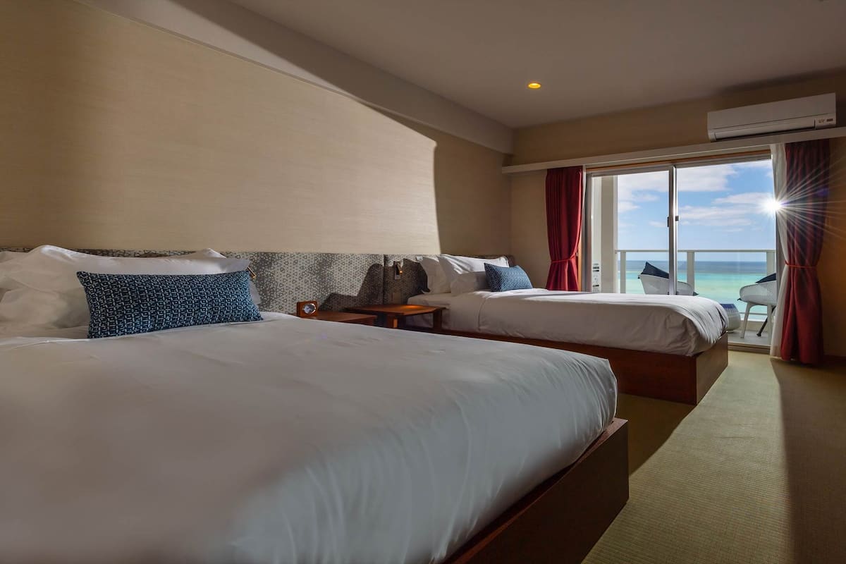 A hotel room with two beds, a TV, a desk, and a balcony with a view of the sea.