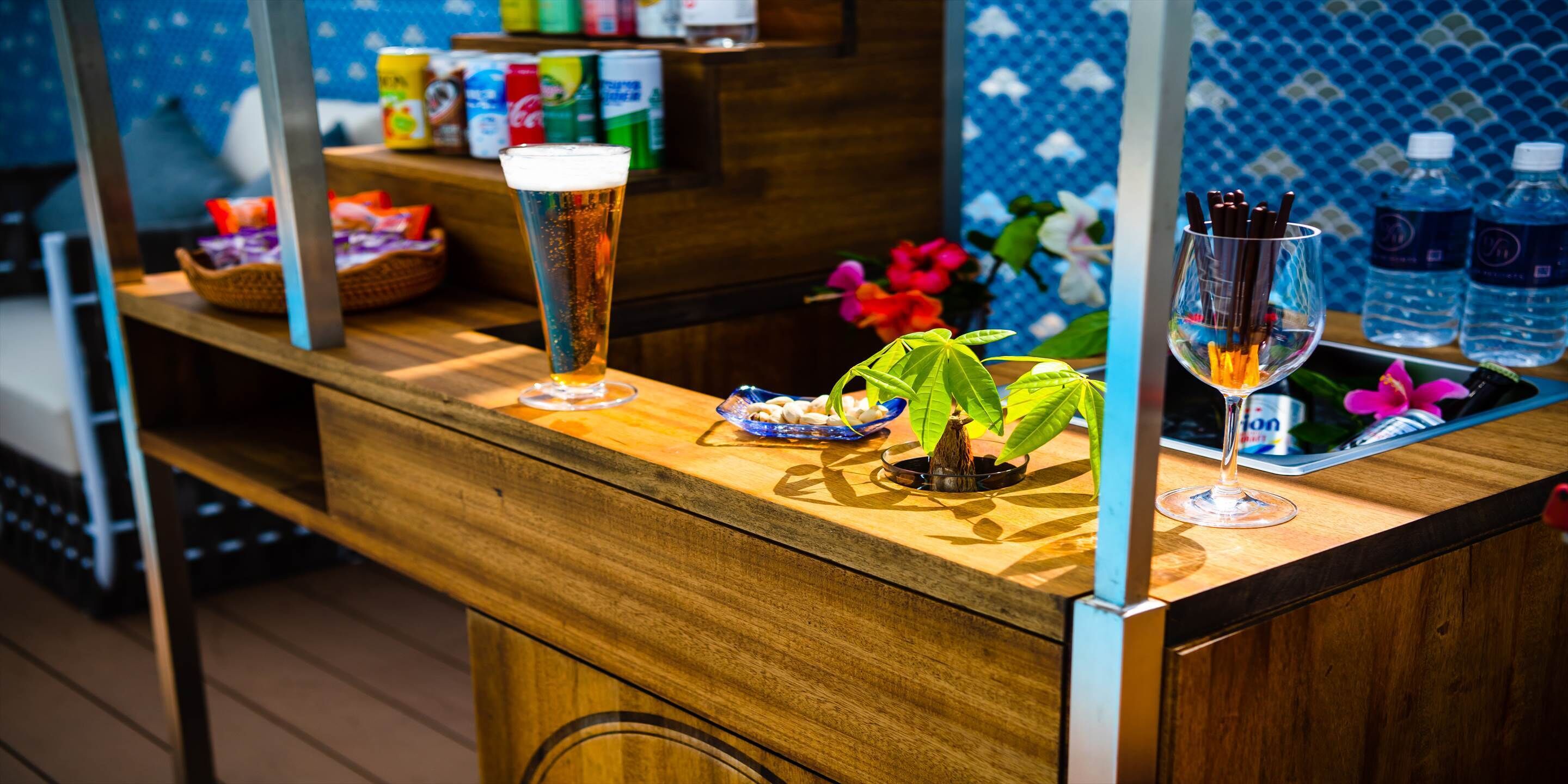 A bar area with a tall glass of beer, a small plant, and a glass with a drink. There are bottles and snacks on the counter.