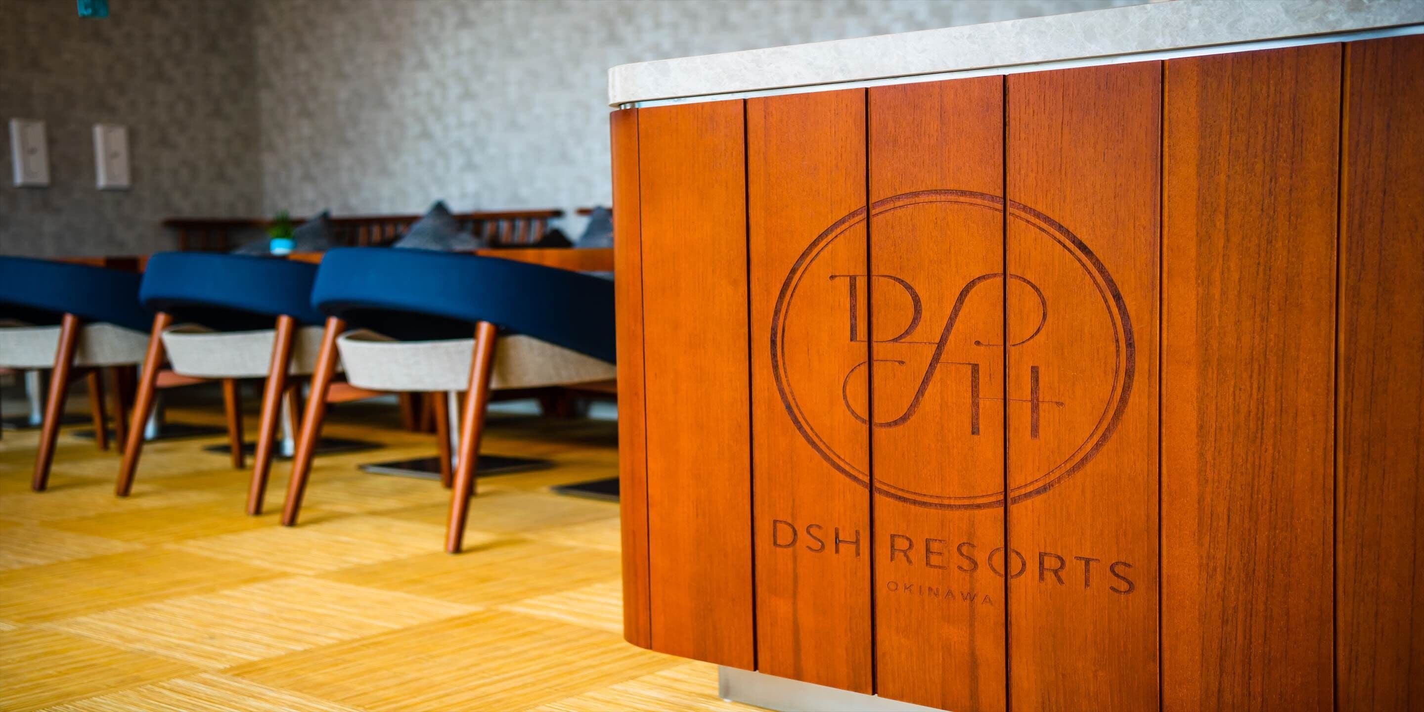 A reception desk with a wooden finish and a logo. The desk is part of a dining area with chairs and a table.