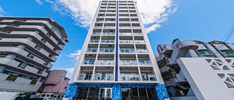 En flereetagers bygning med moderne arkitektur, med altaner på hver etage, en markant indgang med en blå, mønstret facade, og en klar himmel i baggrunden.