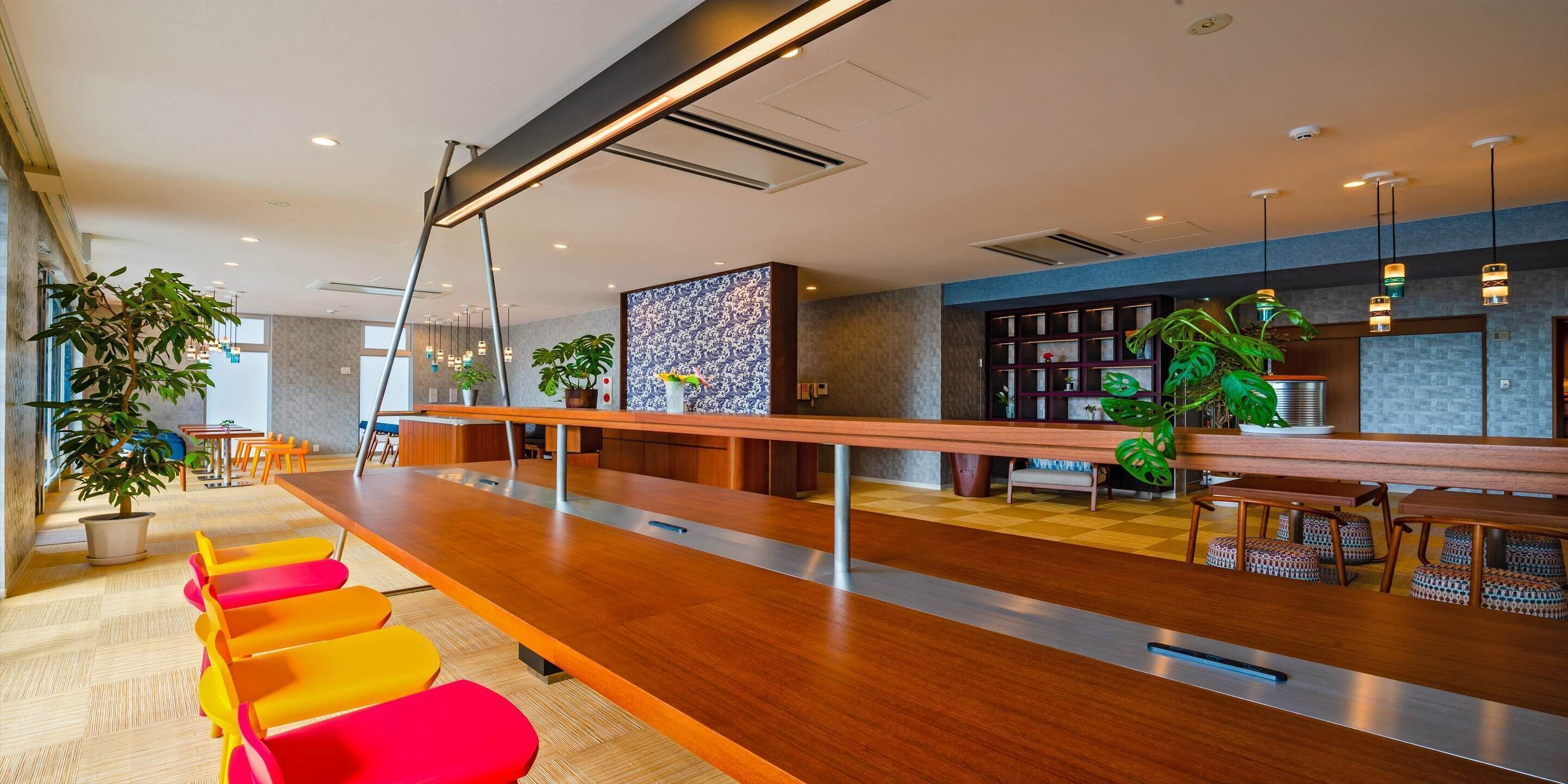 Um saguão moderno de hotel com um balcão de recepção alongado, assentos coloridos, plantas em vasos e uma parede estampada.