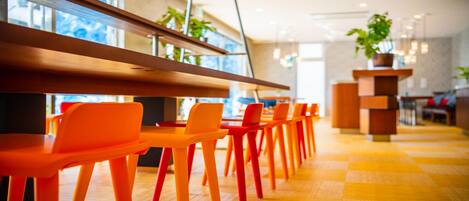 A modern restaurant with orange chairs and wooden tables, large windows with plants, and a checkered floor.