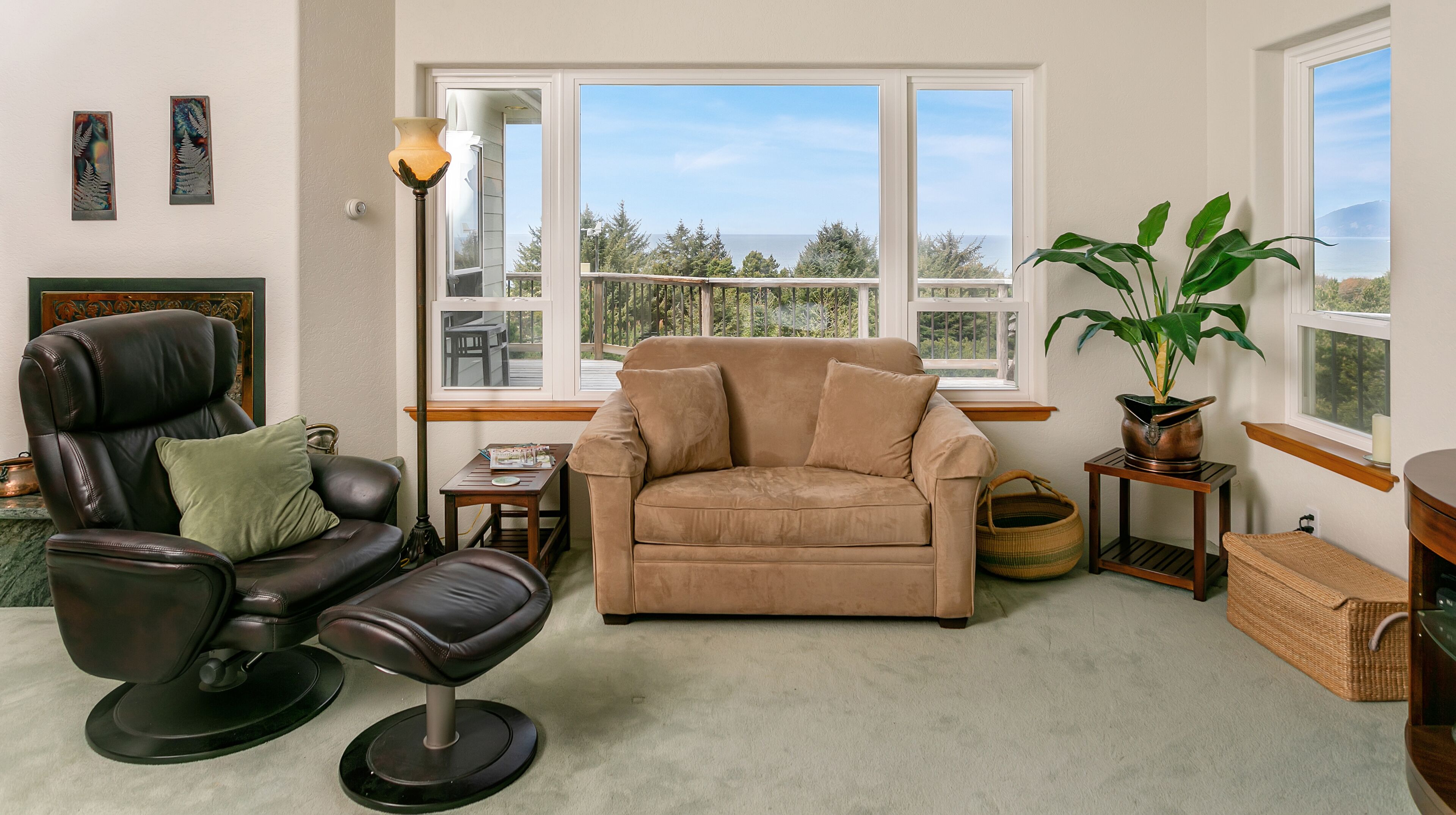 Un salon avec un fauteuil en cuir, un canapé beige, une lampe sur pied et une vue sur l’extérieur grâce à de grandes fenêtres.