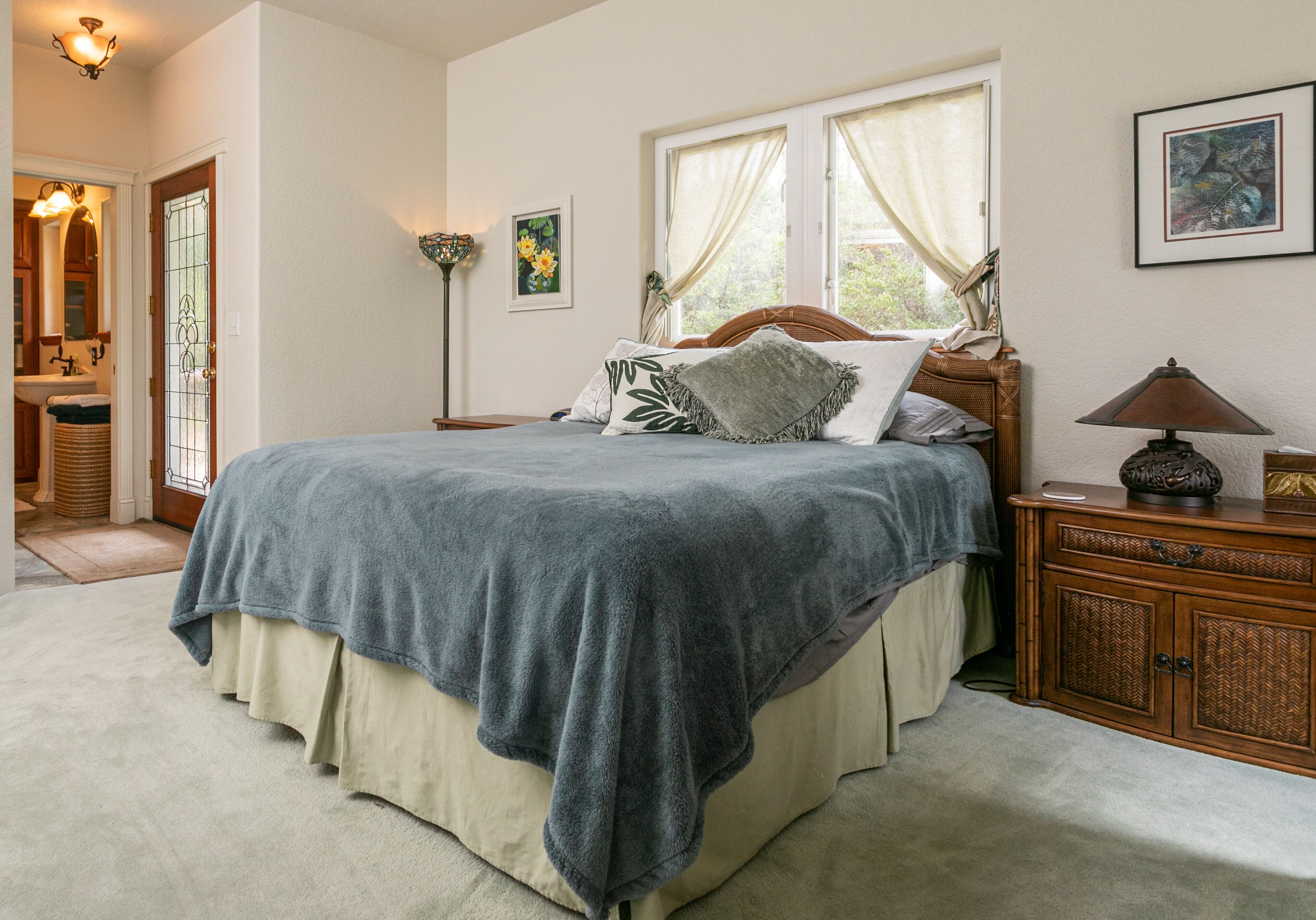 A bedroom with a large bed, a wooden nightstand, a lamp, and a window with curtains.