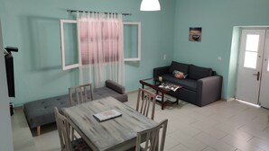 Een woonkamer met een bank, een eettafel met stoelen, een salontafel, een hanglamp, een raam met gordijnen en een deur.