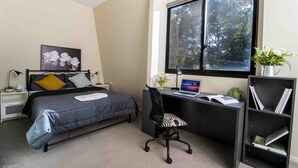 Un dormitorio con cama, escritorio, silla, estantería, ventana y un cuadro.