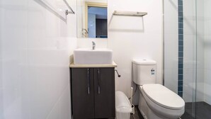 Un baño moderno con lavamanos blanco, mueble de almacenamiento oscuro, inodoro y cabina de ducha con mampara de vidrio.