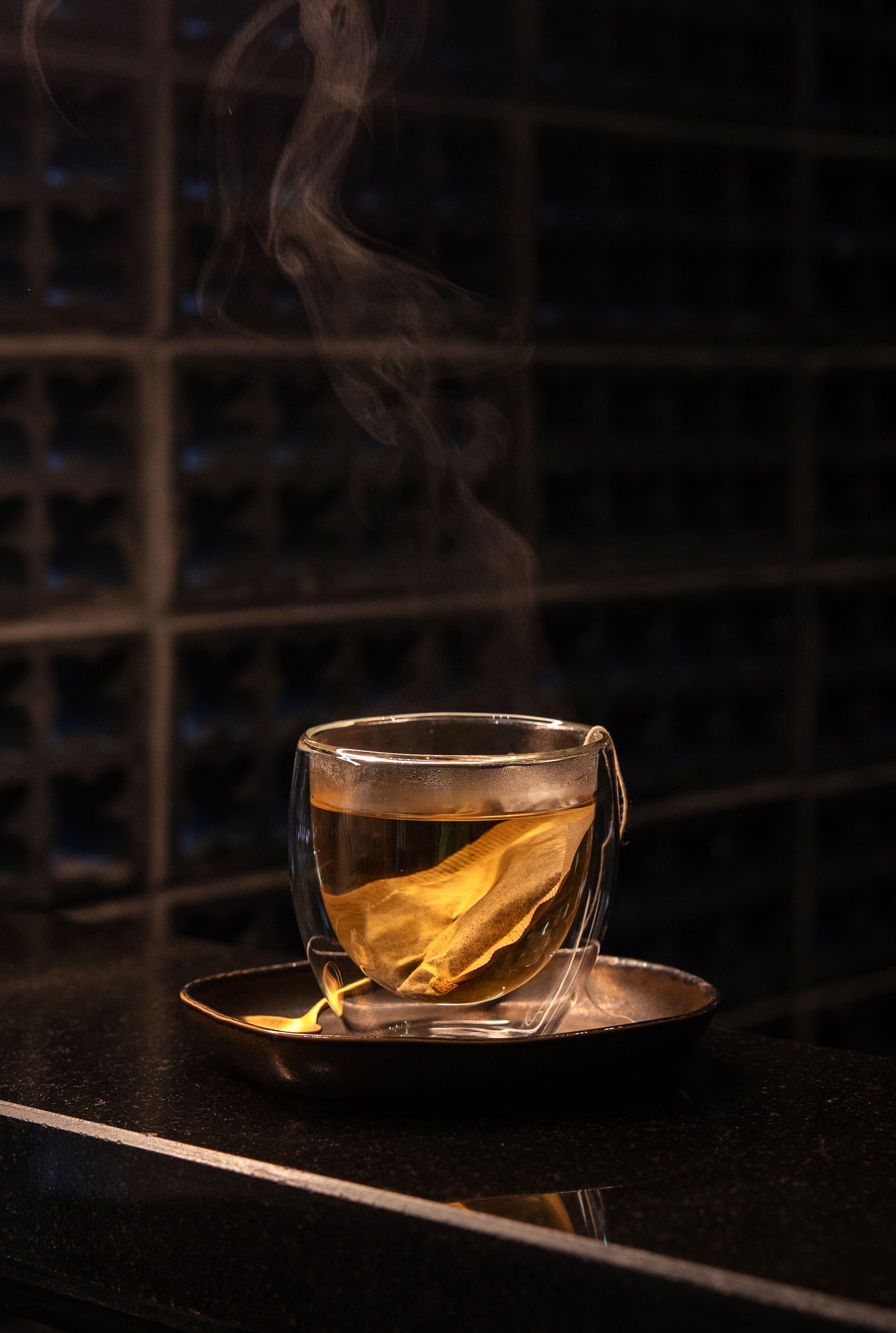 A clear glass cup with a tea bag on a reflective surface, steam rising from it, against a dark background with a grid pattern.