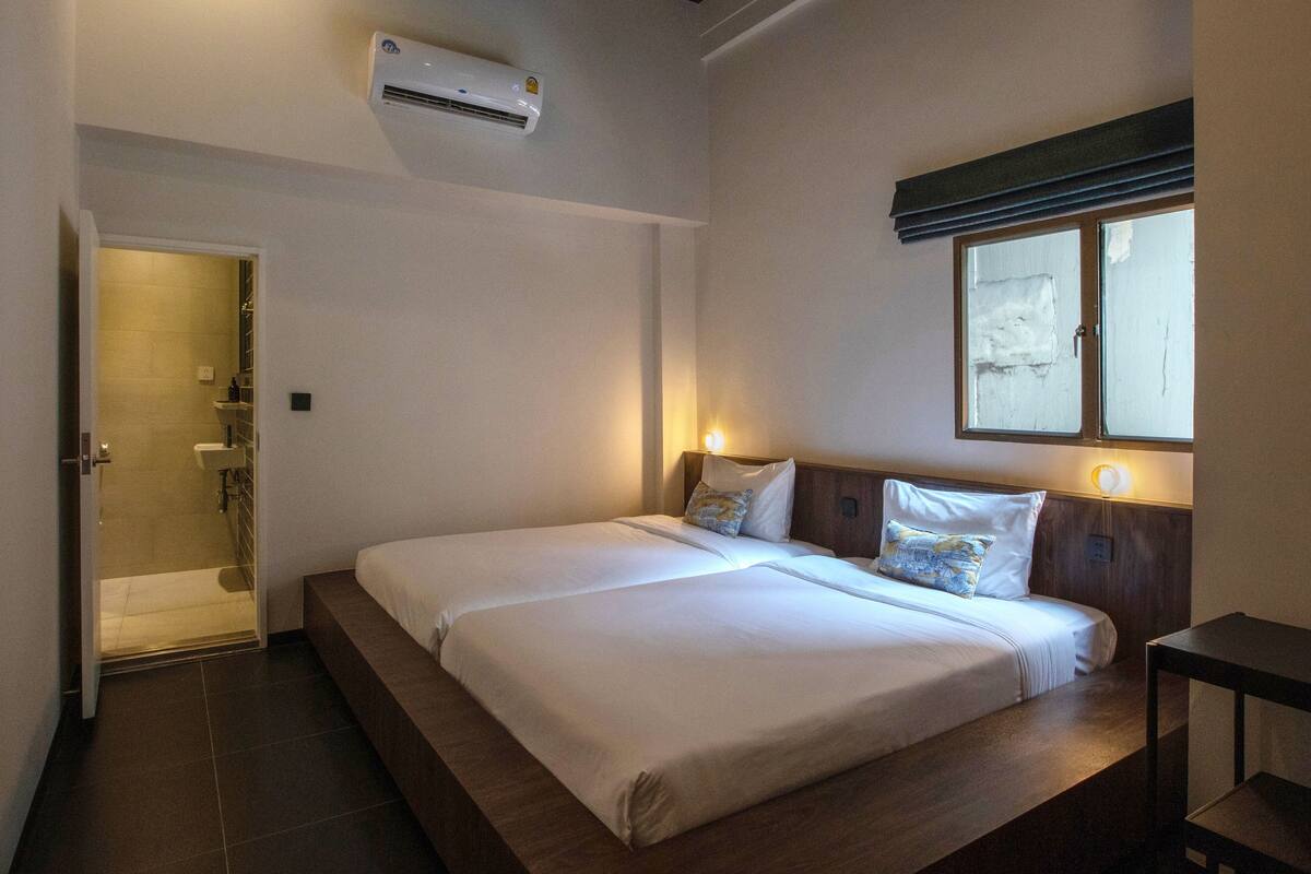 A hotel room with a large bed, a bedside table, a window with blinds, and an air conditioning unit.