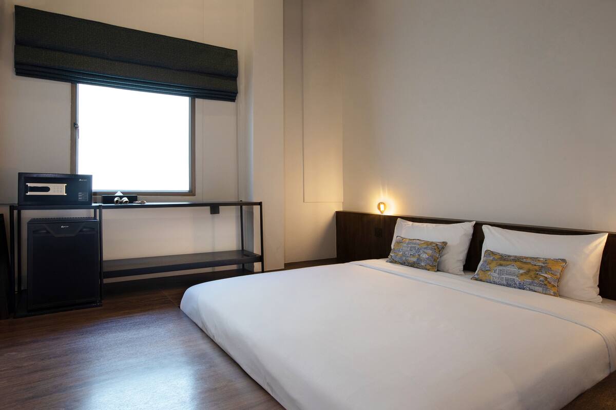 A hotel room with a bed, a desk with a computer, and a window with blinds.