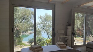 Een slaapkamer met een bed, een stoel en uitzicht op een meer en bomen door grote schuifdeuren.