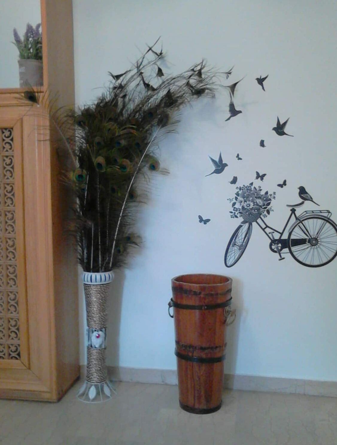 Un vase décoratif orné de plumes de paon et un seau en bois, le tout placé devant un mur orné d’une fresque représentant des oiseaux et un vélo.