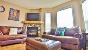 A living room with a stone fireplace, leather sofas, a flat-screen TV, and a painting on the wall. - 3-Story Condo / Mountain Views / Board Games (Fernie)