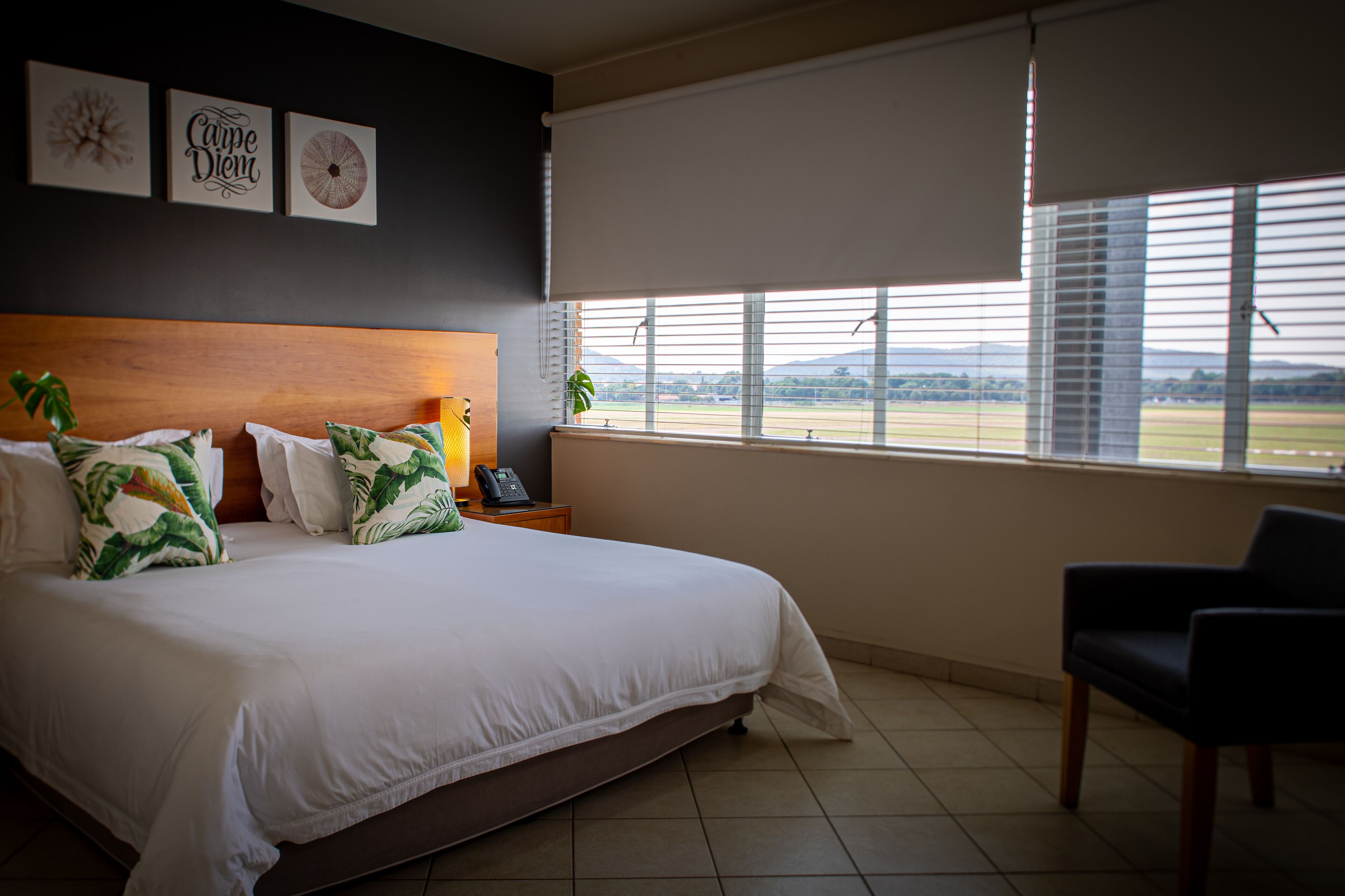 A hotel room with a large bed, a wooden headboard, a nightstand with a lamp and phone, a chair, and a window with blinds.