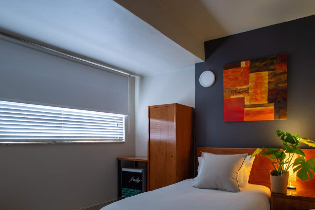 A bedroom with a bed, a wardrobe, a bedside table with a lamp, a window with blinds, and a wall with a painting.