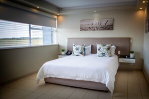 A modern bedroom with a large bed, a bedside table with a lamp, a small plant, and a painting on the wall. - Villa San Giovanni Accommodation (Pretoria)