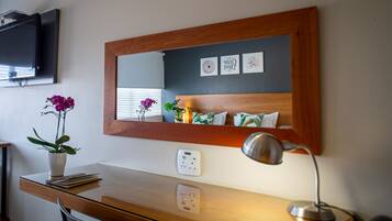 Una habitación de hotel con un escritorio de madera, un espejo, un televisor montado en la pared y una orquídea en maceta.