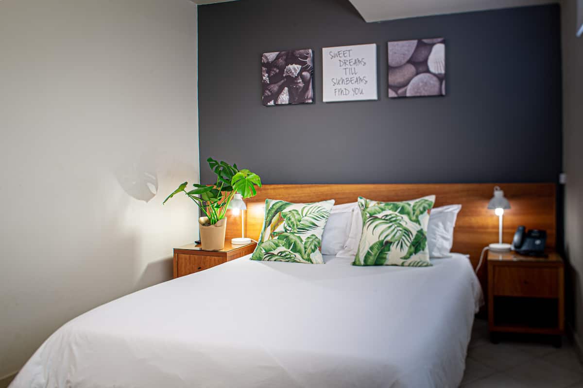 A neatly made bed with white bedding, a wooden headboard, and tropical-themed pillows. There are two bedside tables with lamps and a potted plant. The wall features three framed artworks.