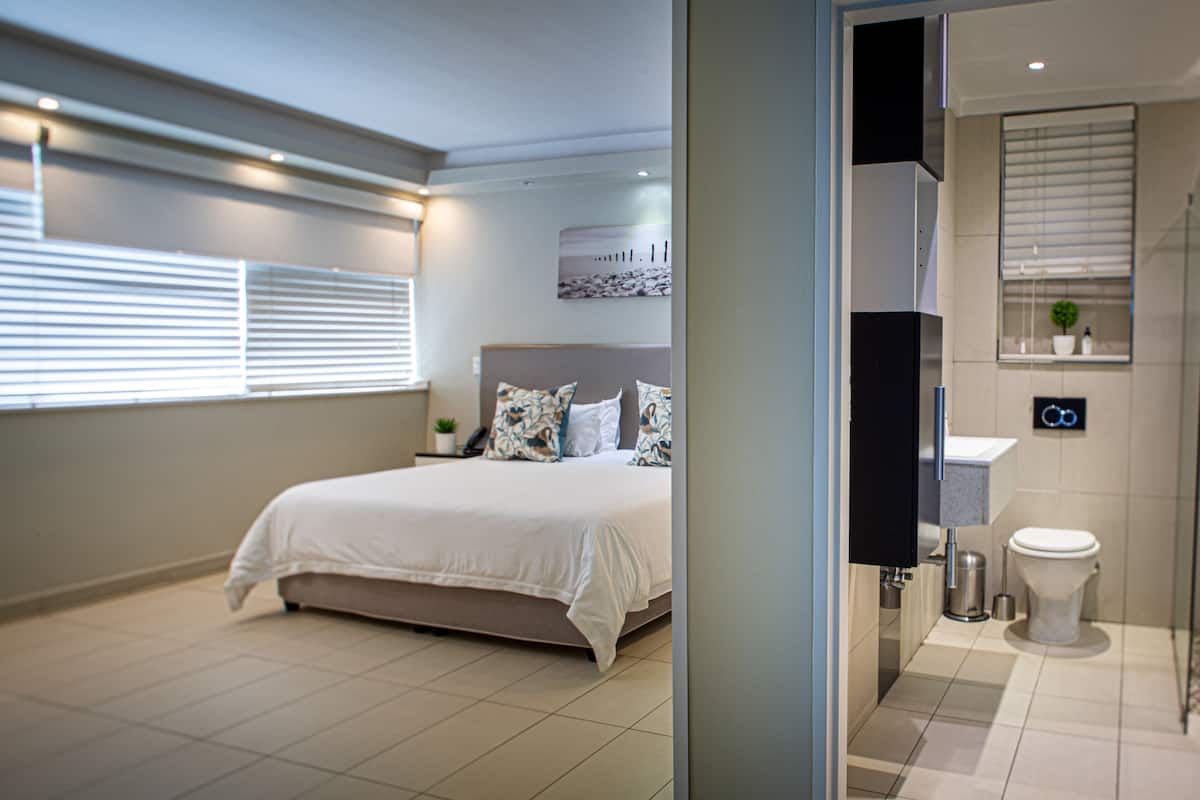 A modern bedroom with a large bed, a bedside table with a plant, and a bathroom with a toilet and sink.