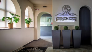 Un espace d’accueil d’hôtel avec une enseigne indiquant « Hébergement près de l’aéroport de Villa San Giovanni ». Des plantes en pot sont disposées sur le rebord de la fenêtre et un paillasson se trouve à l’entrée.