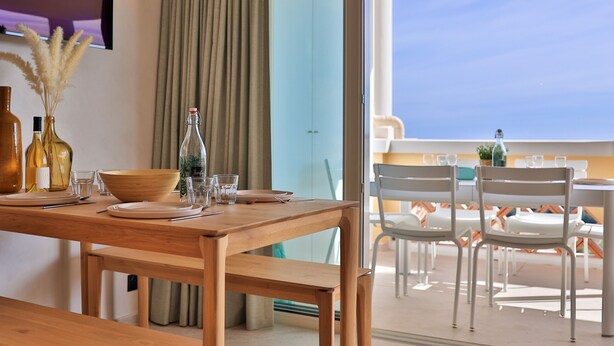 Een houten eettafel gedekt met borden, glazen en flessen, met uitzicht op een balkon en buitenstoelen.