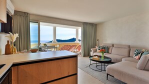 Een moderne hotelkamer met een keukenhoek, een zithoek met een bank en stoelen, en een balkon met uitzicht op zee.