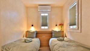 Een kleine, gezellige kamer met twee eenpersoonsbedden, een raam met zonwering, een airconditioning en een klein tafeltje met een bloevas.