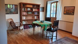 Une pièce avec un plancher en bois, une bibliothèque, une table à manger avec des chaises, un fauteuil en osier, un radiateur et une fenêtre avec des rideaux.