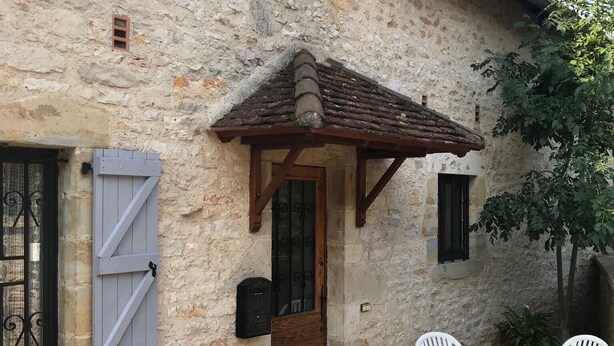 Un bâtiment rustique en pierre, doté d’une porte en bois, d’un petit porche avec deux chaises blanches et d’une plante en pot.