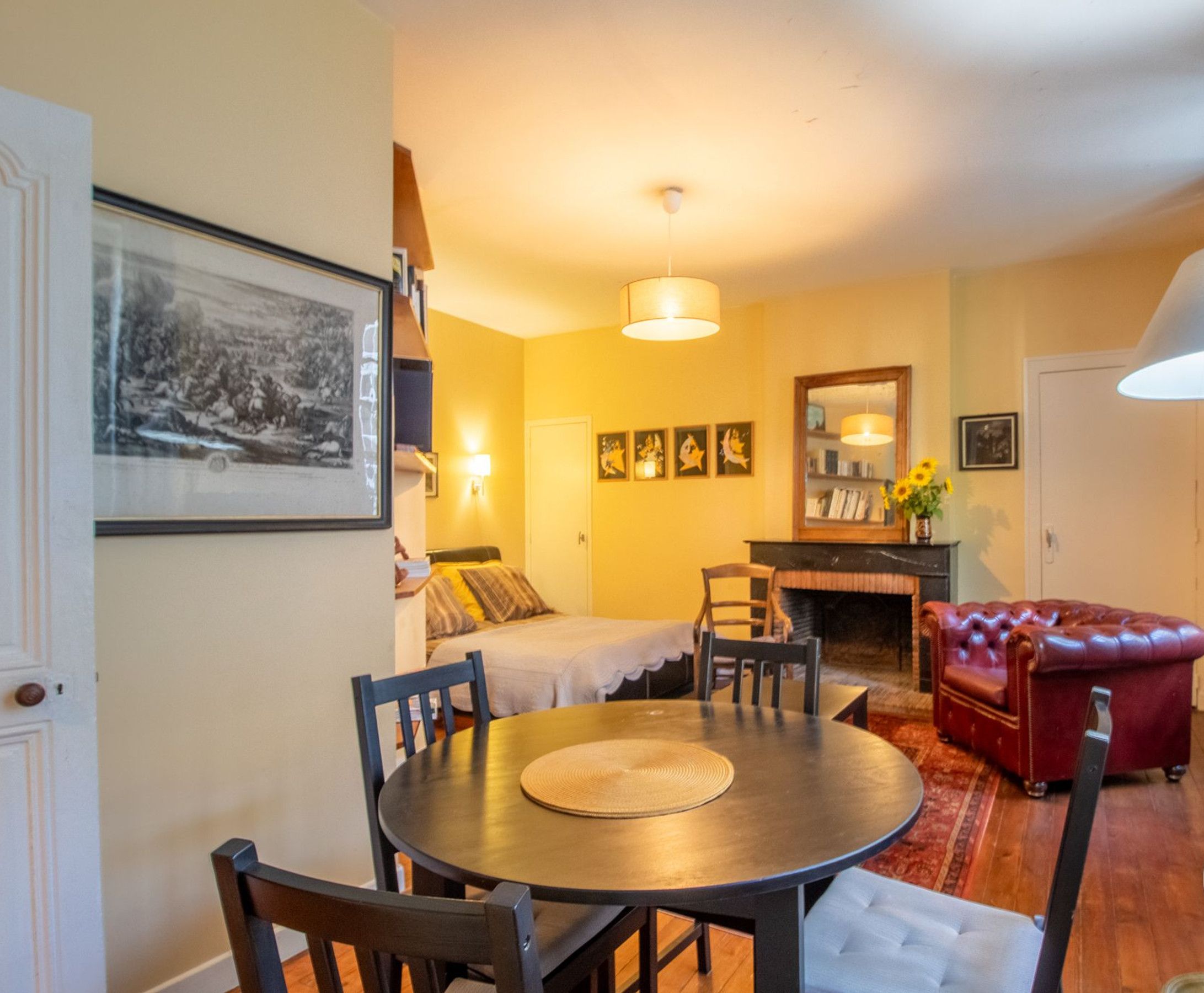 Un salon chaleureux avec une table à manger ronde, un canapé en cuir rouge et une cheminée. Des tableaux encadrés ornent le mur, et une lampe est posée sur une table d’appoint.