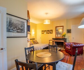 Un salon chaleureux avec une table à manger ronde, un canapé en cuir rouge et une cheminée. Des tableaux encadrés ornent le mur, et une lampe est posée sur une table d’appoint.