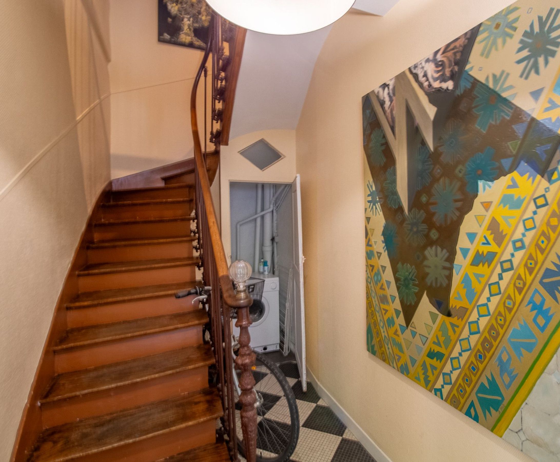 Un escalier en bois avec une rampe, un luminaire fixé au mur et un tableau abstrait aux couleurs vives.