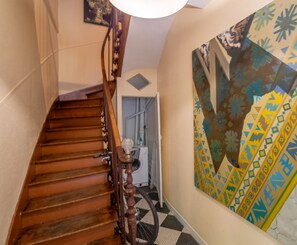 Un escalier en bois avec une rampe, un luminaire fixé au mur et un tableau abstrait aux couleurs vives.