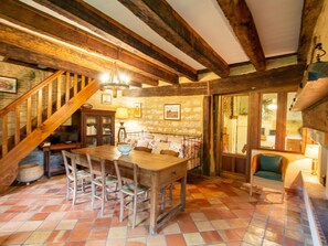 A rustic interior with exposed wooden beams, a tiled floor, a wooden dining table with chairs, a staircase, and a window with curtains.