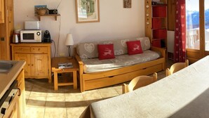 A cozy room with a wooden sofa, a small table, a kitchenette, and a view of the outdoors.
