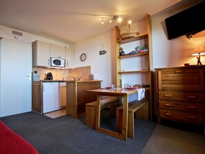 A compact kitchen with a microwave, sink, and wooden cabinets. There is a dining table with chairs and a small bench. A television is mounted on the wall, and a clock is visible.