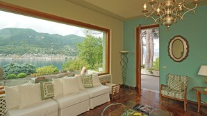 A living room with a sofa, a chair, a table, a chandelier, and a large window offering a view of a lake and mountains.
