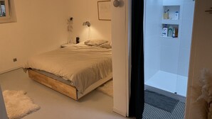 A bedroom with a bed, a bedside lamp, a small table, and a door leading to a bathroom with a shower and shelves with toiletries. - Maison de Charme Marseille Borely (Marseille)