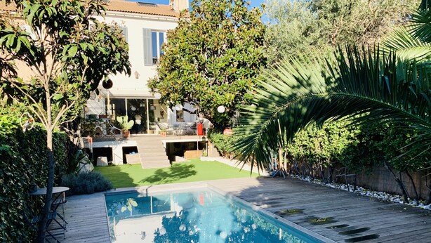 A swimming pool with a wooden deck, surrounded by lush greenery and a house in the background. - Maison de Charme Marseille Borely (Marseille)