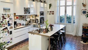 A modern kitchen with white cabinets, a central island with stools, a hanging pot rack, and a large window with curtains. - Maison de Charme Marseille Borely (Marseille)