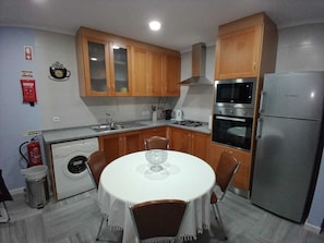 A compact kitchen with wooden cabinets, a stainless steel refrigerator, a built-in oven, and a washing machine. - Feather House Home (São Bartolomeu de Galegos)