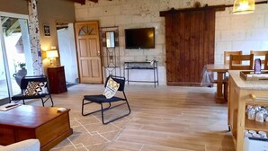 A spacious living area with a wooden floor, a television mounted on the wall, and a dining area with a table and chairs. - Charming Cottage near Loire River with Garden, Covered Terrace & Easy Access to Saumur Attractions (Brain-sur-Allonnes)