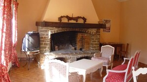 Uma sala de estar acolhedora com lareira de pedra, televisão sobre um móvel e um espaço de jantar com cadeiras brancas e vermelhas.