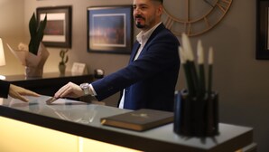 A man in a blue suit standing at a reception desk with framed pictures on the wall behind him. - Hotel Opera (Pristina)