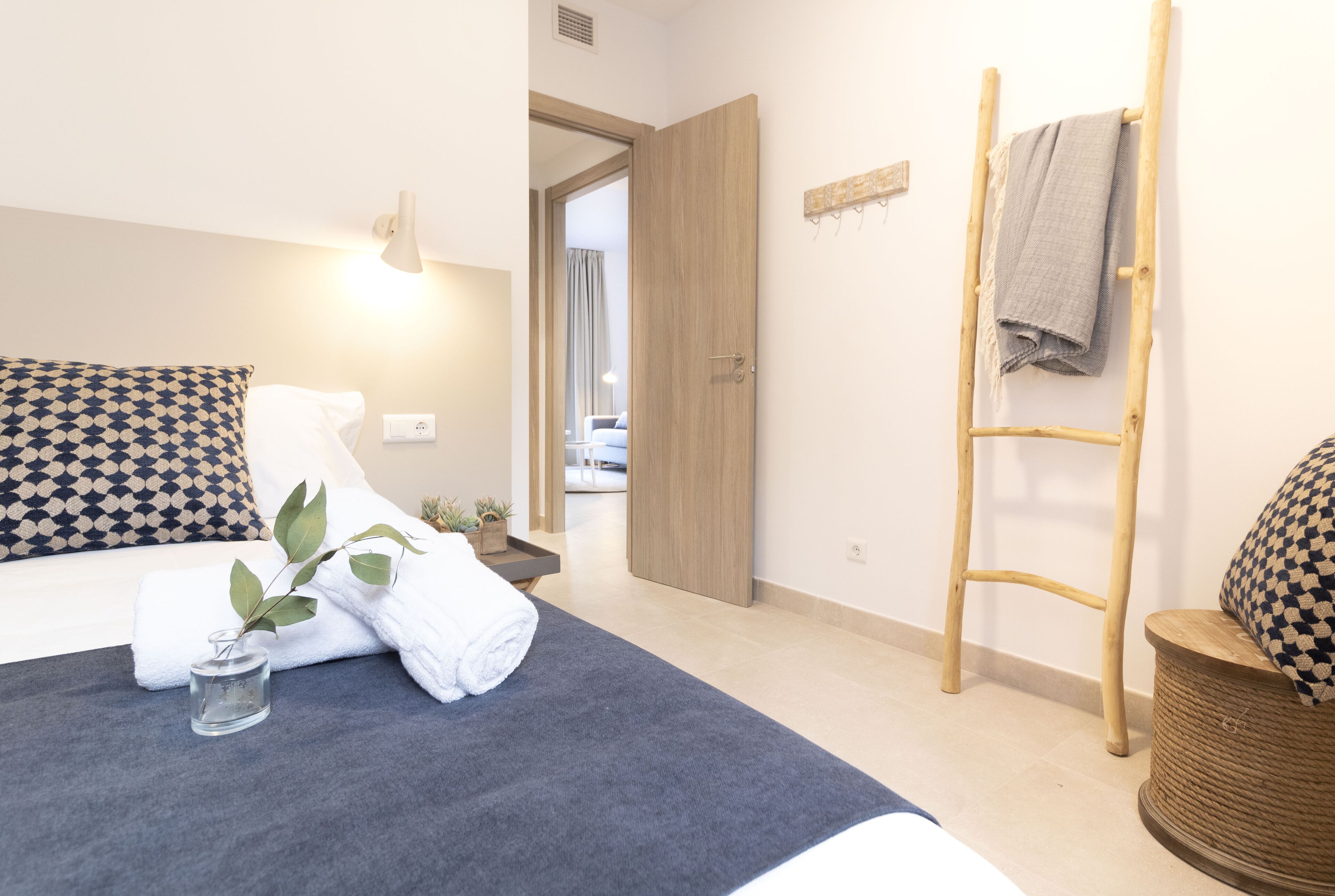 Hotelkamer met een bed, nachtkastje met een vaas, handdoek, houten ladder, en zicht op de aangrenzende kamer.