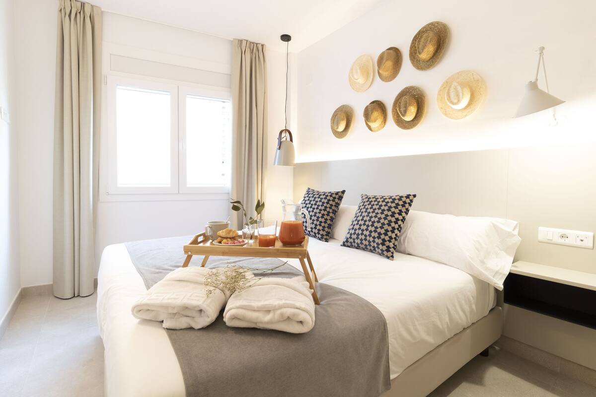 Una cama bien tendida con una bandeja de desayuno, una mesita de noche y una pared decorada con sombreros.