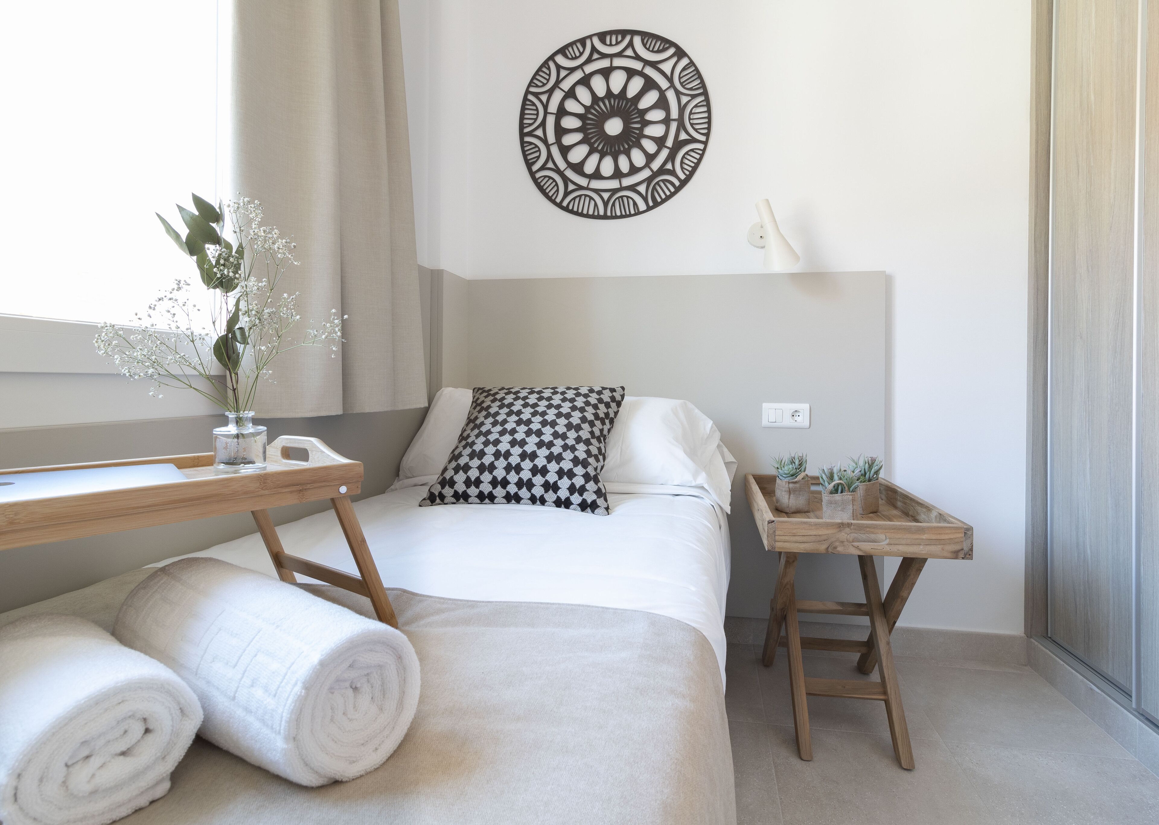 A neatly arranged bedroom with a bed, a bedside table with a lamp, a decorative wall piece, and a small plant on a wooden side table.