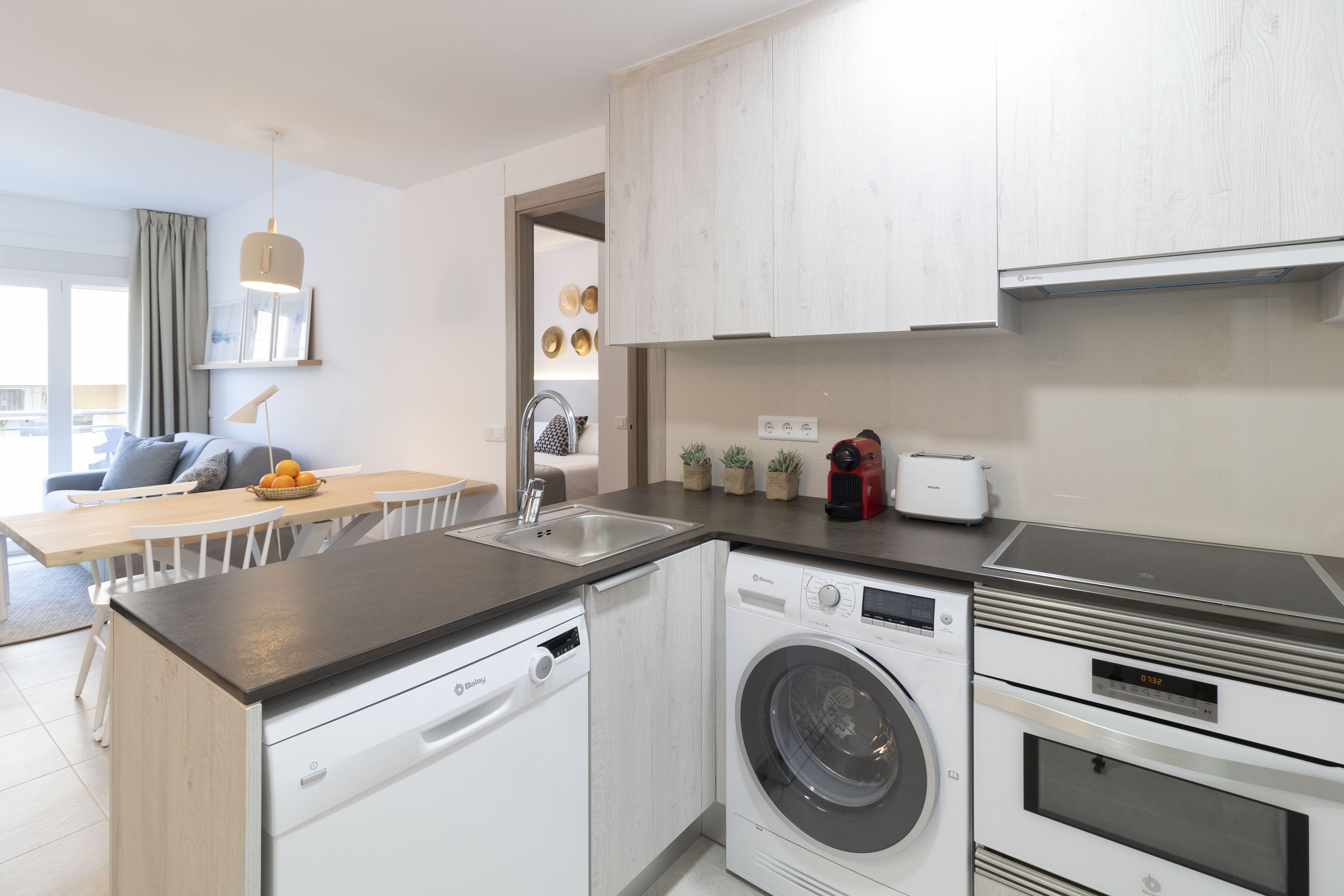 A modern kitchen with a dishwasher, washing machine, and oven. There is a dining area with a table and chairs.