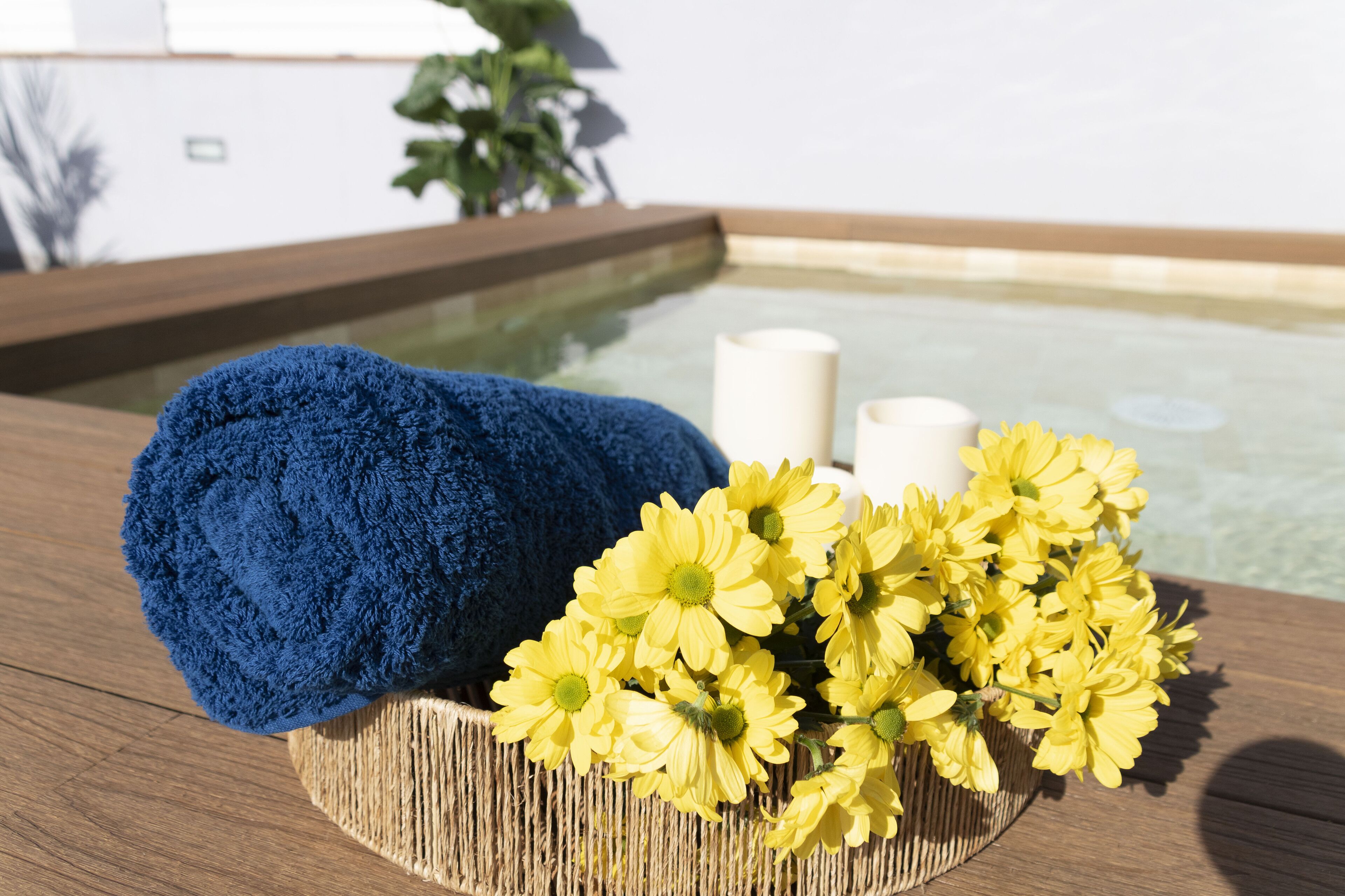 Une table en bois sur laquelle est posée une serviette bleue, un panier en osier rempli de fleurs jaunes et une bougie blanche.